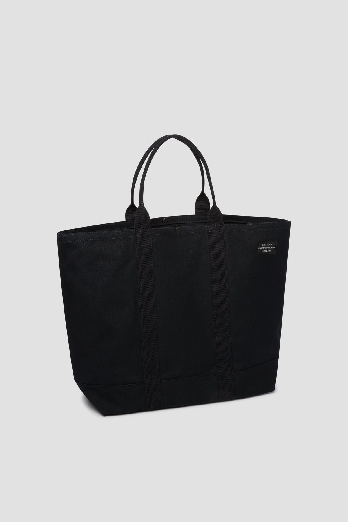 Black canvas tote bag with top button, short handles, and front patch logo.