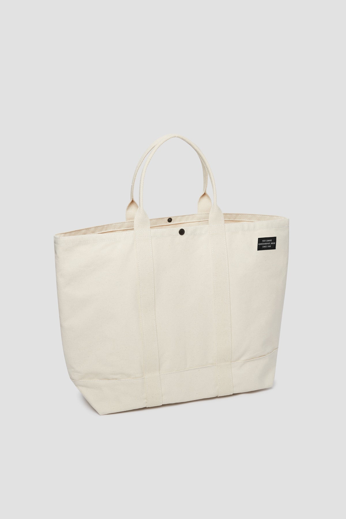 Natural canvas tote bag with top button, short handles, and front patch logo.